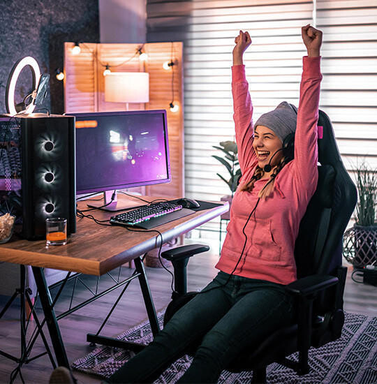 Person in rosa Kapuzenpullover sitzt in Bürostuhl, streckt Arme nach oben, vor Schreibtisch mit Computer und Tastatur.